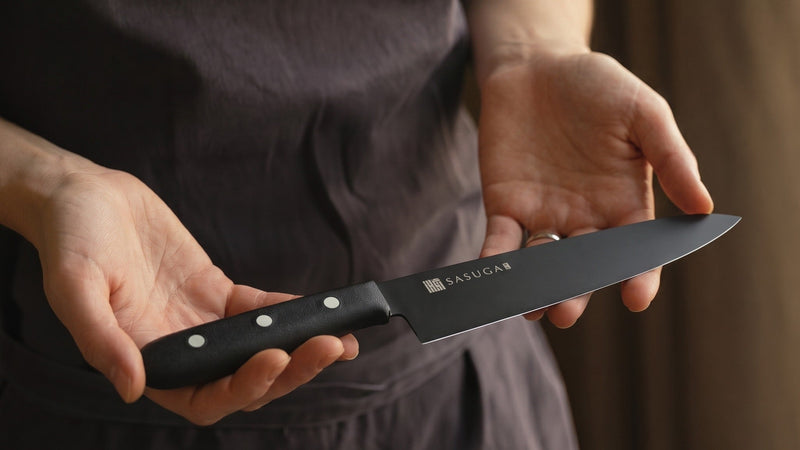 Hands presenting a SASUGA Gyuto Japanese chef's knife with black blade